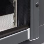 Lähikuva Hyttysverkko ikkunaan - rullaverkko ikkunaruudun nurkasta, jossa on harmaa kehys ja valkoinen muovinen liitospala.