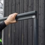 Joku asentaa Hyttysverkko ikkunaan - rullaverkko kehyksen tummaa puuseinää vasten.