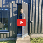 Mies seisoo oviaukossa Hyttysverkko oveen - avattava auki, ulospäin; vasemmalla tumma puuseinä ja ikkuna.