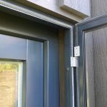 Lähikuva tummasta karmista, jossa on avattu Hyttysverkko oveen - avattava, jossa näkyy sarana ja osa seinän sivuraidetta.