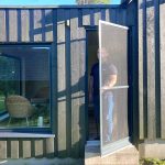 Mies seisoo Hyttysverkko oveen - avattavan takana tumman puutalon sisäänkäynnillä, auringonvalo heittää varjoja.