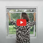 A woman installs an Insect screen window – roller blind in a white frame, with green trees outside.