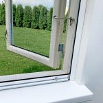 Avoin valkopuitteinen ikkuna Hyttysverkko ikkunaan - rullaverkko, josta näkymät vihreälle nurmikolle ja korkeille puille.
