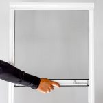 Käsi vetää alas Hyttysverkko ikkunaan - rullaverkko ikkunaseinää vaaleaa taustaa vasten.