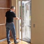 Ein Mann in Freizeitkleidung öffnet ein Fliegengitter Tür - faltbar, um aus einem sonnenbeschienenen Raum nach draußen zu treten.