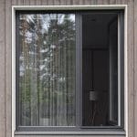 Hyttysverkko ikkunaan - kehysmalli, jossa on musta kehys, on osittain auki ja heijastaa ulkona olevia puita; sisällä näkyy lamppu.