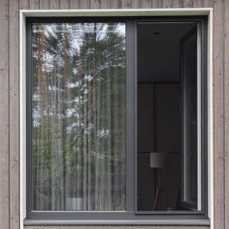 Hyttysverkko ikkunaan - kehysmalli, jossa on musta kehys, on osittain auki ja heijastaa ulkona olevia puita; sisällä näkyy lamppu.