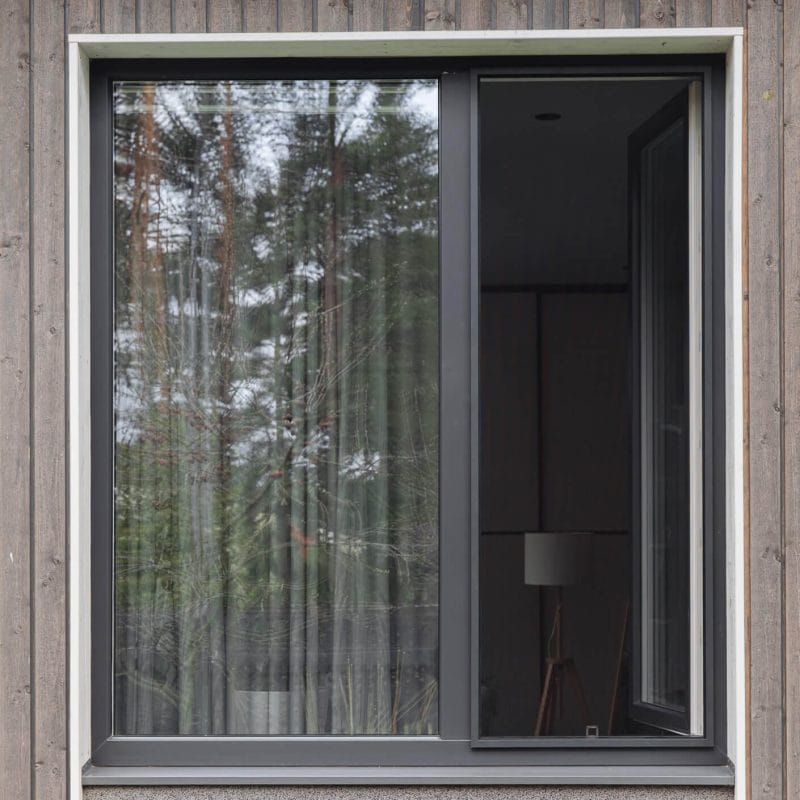 Hyttysverkko ikkunaan - kehysmalli, jossa on musta kehys, on osittain auki ja heijastaa ulkona olevia puita; sisällä näkyy lamppu.