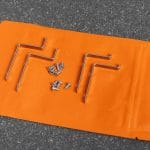 Three metal L-brackets and screws for the Insect screen window – in frame are displayed on an orange surface with a gray backdrop.