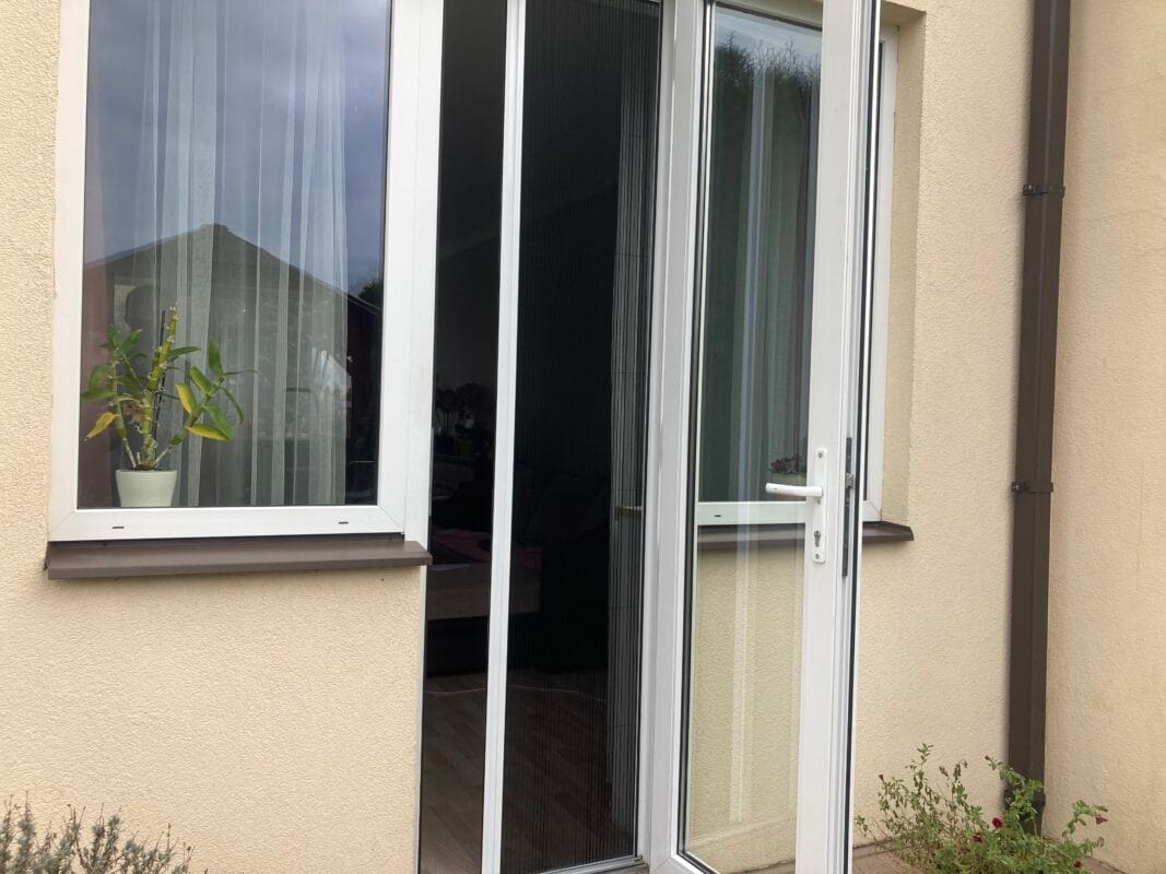 Eine teilweise geöffnete Glastür und ein daneben liegendes Fenster mit insektu sieti an einem beigen Gebäude, dazu eine Topfpflanze auf der Fensterbank.