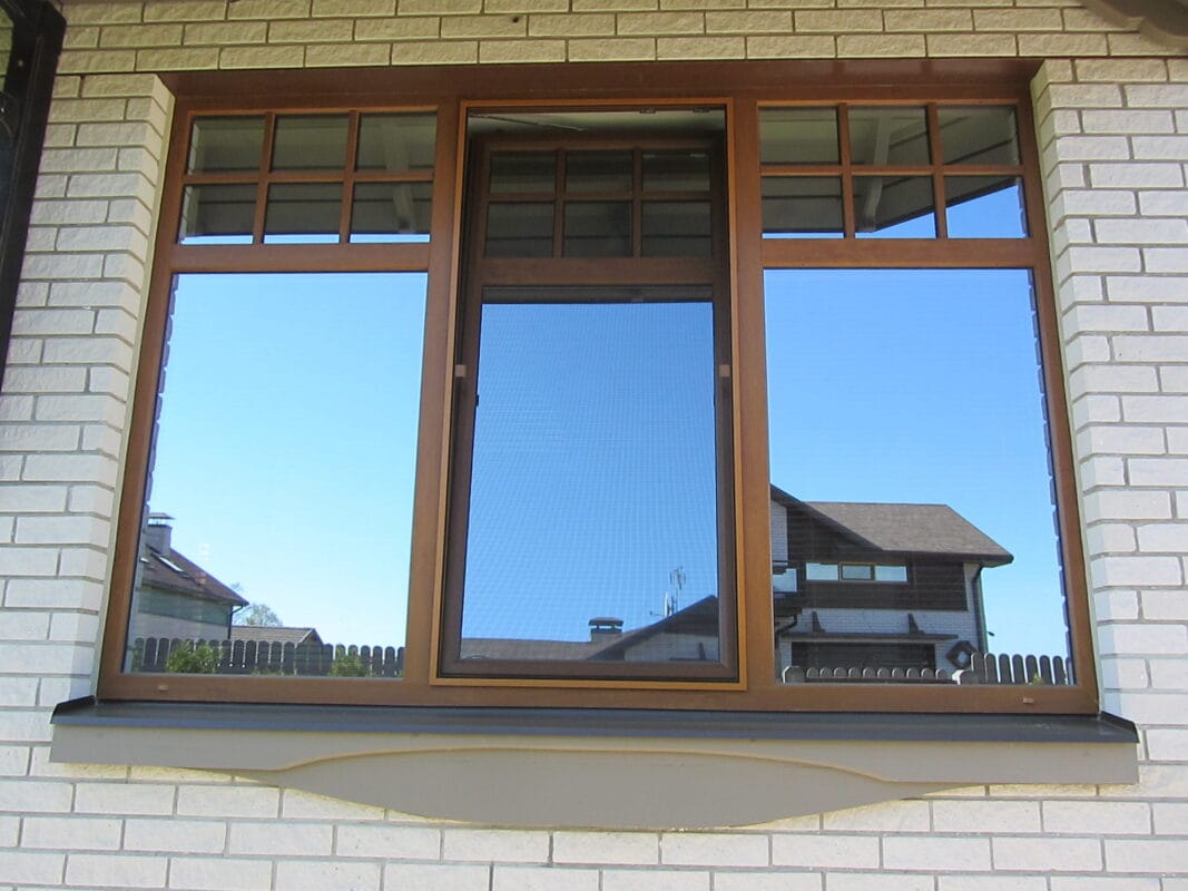 Ein großes Fenster mit Holzrahmen und drei Glasscheiben spiegelt ein Haus und den blauen Himmel in einer weißen Backsteinmauer wider.