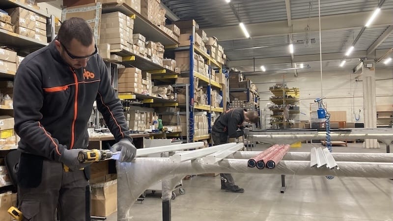 Divi strādnieki montē iekārtas insektu sietu ražošanai noliktavā, kuru ieskauj plaukti ar kastēm un materiāliem.